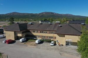 En stor tvåvånings tegelbyggnad med platt tak och flera parkerade fordon framför, mot en bakgrund av gröna träd och avlägsna berg under en klarblå himmel.